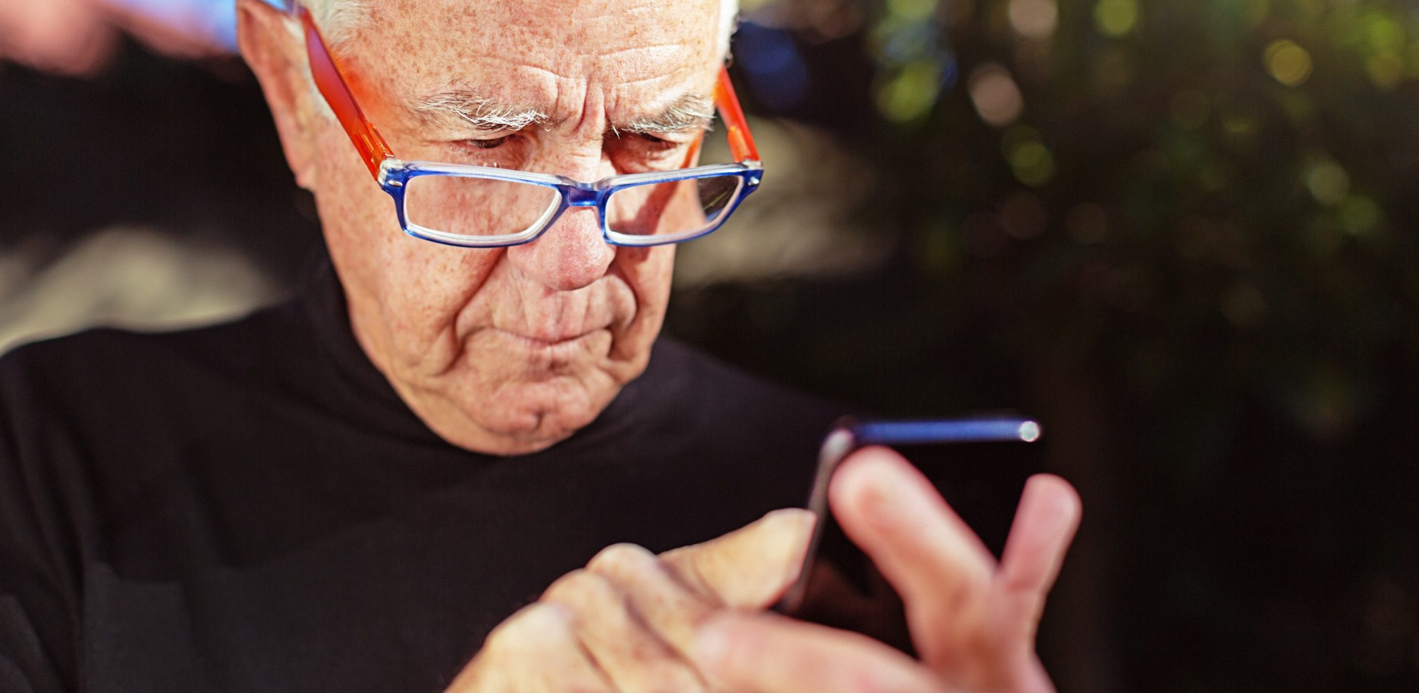 Senior man squinting over rim of glasses at phone.